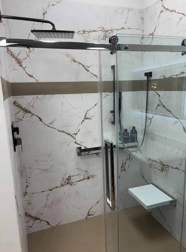Remodeled walk-in shower with gold-veined marble-look walls, black rainfall shower head, glass doors and fold-down seat